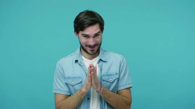 Sneaky scheming bearded guy in jeans shirt rubbing hands and plotting cunning idea, smirking and conspiring villain plan, looking with greedy sly expression. studio shot isolated on blue background