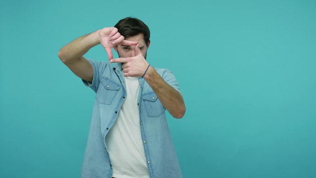 Cameraman cropping picture. Bearded guy in jeans shirt looking through photo frame made of hands, zooming distance, focusing at camera, capturing moment. indoor studio shot isolated on blue background