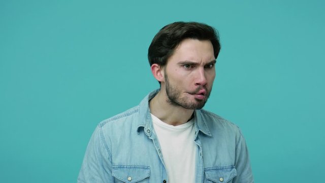 Comical weird bearded guy in jeans shirt watching around making fish face with pout lips, frowning and looking with humorous wary suspicious expression. indoor studio shot isolated on blue background