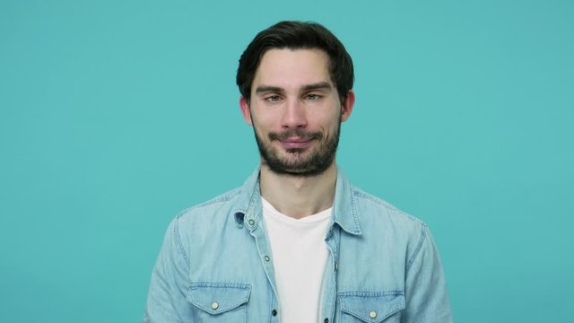 Comical stupid bearded guy in jeans shirt looking cross-eyed and making funny awkward silly grimace, playing fool, pretending to be dumb, brainless. indoor studio shot isolated on blue background