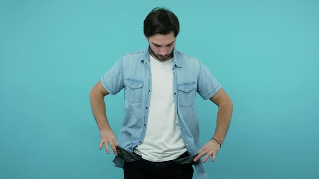 Poor student, jobless bearded guy in jeans shirt worried of overspend, showing empty pants pockets having no money, concept of poverty bankruptcy debts. indoor studio shot isolated on blue background