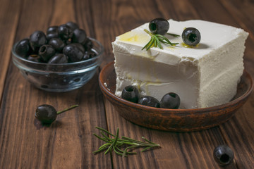 A bowl of black olives and a large piece of feta cheese on a wooden table.