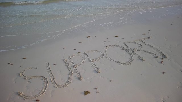 Angled shot of SUPPORT written in the sand on a beach with water lapping onto the sand.
