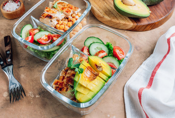 Healthy meal prep containers with chickpeas, chicken, tomatoes, cucumbers and avocados. Healthy lunch in glass containers on beige rustic background. Zero waste concept. Selective focus.