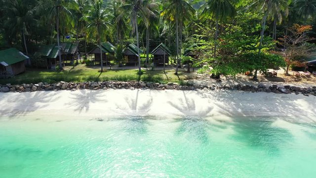 Beach cabins under palm trees with green leaves shade over white sand of exotic beach washed by calm turquoise lagoon