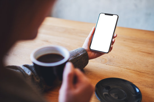 Mockup image of a woman holding  mobile phone with blank white screen while drinking coffee on wooden table