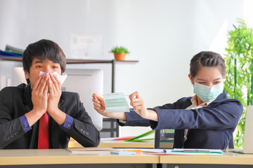 Female employee giving medical face mask to her colleague who is sneezing while working in business office for preventing germs spreading