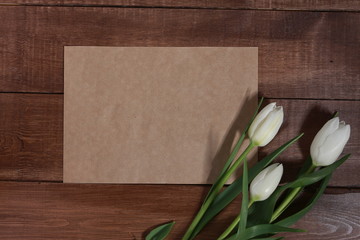 White tulips and blank sheet of craft paper for message or text on brown wooden table. Spring concept. 8 march, women or mothers day, flowers