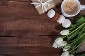 Fototapeta premium Bunch of white tulips, gifts, macaroons and cup of coffee on brown wooden table. Greeting concept. Spring, women or mothers day, cakes, cappuccino