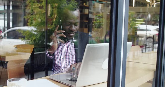 Through cafe window view of happy African businessman talking with clients on the mobile phone looking at laptop on the table checking information urban street reflection on the window glass