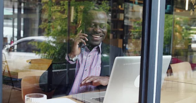 Through cafe window view of happy African businessman talking with clients on the mobile phone with laptop on the table urban street reflection on the window glass