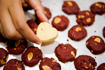 A hand hold a love shape cookies in the middle of red velvet cookies on a tray