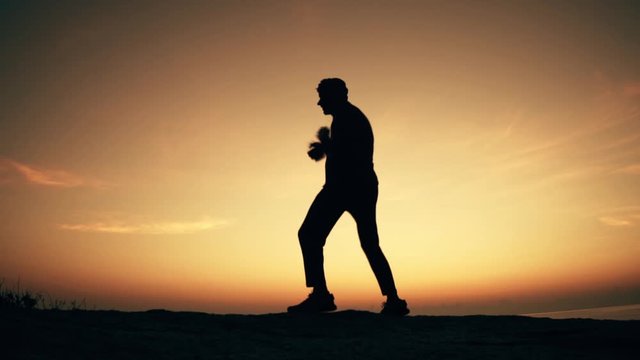 Man practices shadowboxing on a rock by the sea at dawn. Slow motion video.