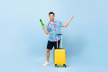 Smiling handsome Asian tourist man traveling with water gun and baggage during Songkran festival studio shot blue background
