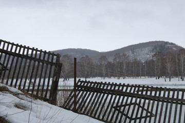 Destroyed old fence against the background of trees and snow -capped mountains.