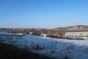 Rural houses on the background of snow covered fields trees and mountains under a blue sky.