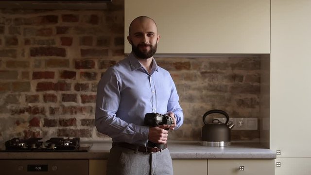 A bald photographer searches for some preferences in his camera then looks straight at the camera with a smile in the dining room. A man with a beard wears two silver earrings and a blue shirt.
