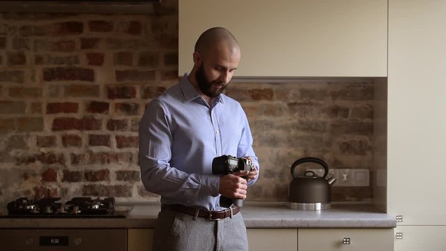 A bald photographer searches for something in his camera in the dining room. A man with a beard wears two silver earrings, a blue shirt, grey trousers, and a brown leather belt.