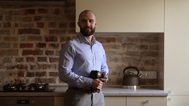 A bald photographer poses with his camera and looks straight with a smile. A man with a beard wears two silver earrings and a blue shirt in the dining room.