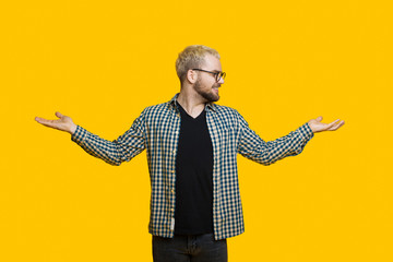 Bearded caucasian man with blonde hair is comparing two things from his palms while posing on a yellow wall