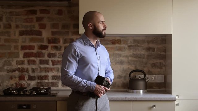 A happy bald photographer with his camera turns his face to the side with a smile. A man with a beard wears two silver earrings and a blue shirt in the dining room.