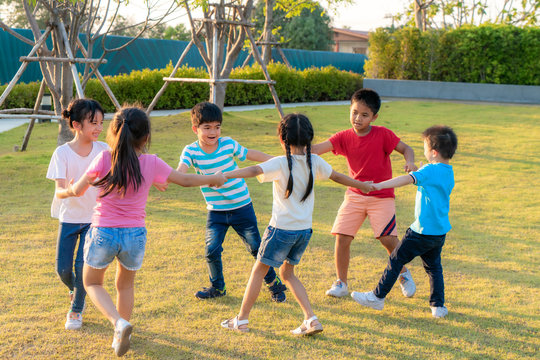 Large group of happy Asian smiling kindergarten kids friends holding hands playing and dancing play roundelay and stand in circle in the park on the green grass on sunny summer day..