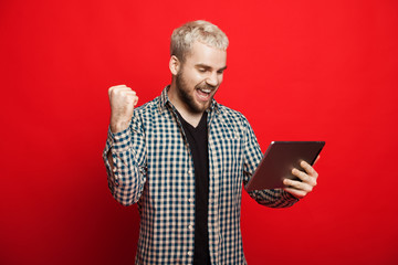 Caucasian man with blonde hair and beard is gesturing a winning while holding a tablet and posing on a red background