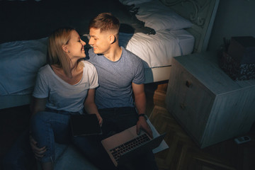 Charming caucasian couple kissing each other while sitting on the floor and using a tablet and laptop