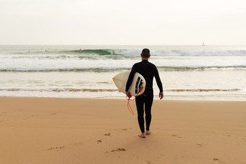 Back view of man with surfboard on beach. Rear view of surfer in wetsuit holding board and walking to sea waves at summertime. Surfing concept
