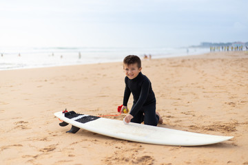 Adorable child waxing surfboard on beach. Cute happy little boy in wetsuit kneeling on sand and waxing surfboard on sea coast at summertime. Surfing concept