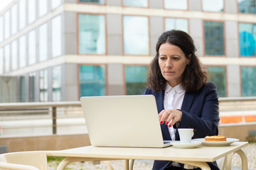 Focused businesswoman using laptop in outdoor cafe. Serious middle aged businesswoman working with laptop computer while sitting at table with coffee outdoors. Technology concept