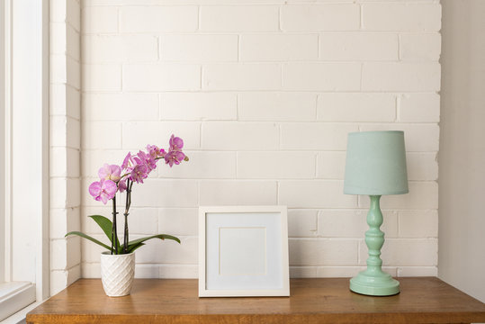 Closeup of purple moth orchid, blank square white picture frame and teal lamp on wooden table against painted brick wall