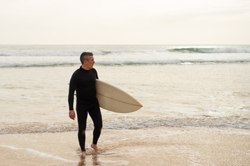 Man holding surfboard and looking at ocean. Full length view of handsome middle aged surfer in wetsuit holding board and looking at waves. Surfing concept