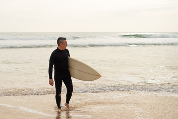 Man holding surfboard and looking at ocean. Full length view of handsome middle aged surfer in...