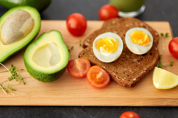 food, eating and breakfast concept - toast bread with eggs, cherry tomatoes, avocado and greens on wooden cutting board