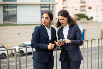 Concentrated businesswomen using smartphone. Female colleagues in formal wear standing on street and using cell phone together. Business and technology concept