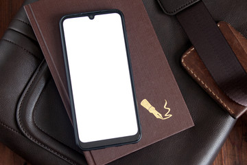 A smartphone with a blank screen on top of a notebook lies on a leather bag on a wooden table.