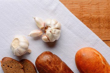 Different kinds of bread with fresh raw garlic on white tablecloth over light rustic wooden table.