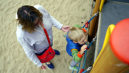 Fototapeta na wymiar Dziecko wspina się po linach na placu zabaw, mama pomaga mu i podtrzymuje, żeby nie spadł.
