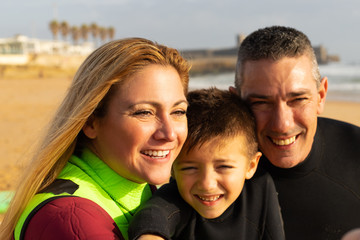 Fototapeta premium Happy family in wetsuits on beach. Close-up portrait of cheerful middle aged parents and cute little son in wetsuits smiling at camera on sea coast. Water sport concept