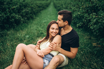 Woman with long hair and man sitting between currant bushes, hugging and kissing. Background of green currant plantations