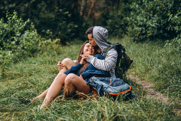Naklejka na ściany i meble Woman with long hair and man sitting on the grass in the forest with backpacks. Have fun in nature, hugging and kissing