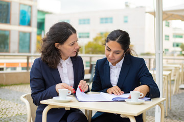 Fototapeta premium Businesswomen working with papers in outdoor cafe. Professional female colleagues in formal wear sitting at table, drinking coffee and discussing work in outdoor cafe. Coffee time concept