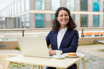 Fototapeta na wymiar Businesswoman using laptop and smiling at camera. Happy middle aged businesswoman working with laptop computer while sitting at table with coffee outdoors. Technology concept