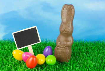 Colorful easter eggs on a meadow with a slate blackboard and a chocolate Easter bunny, blue sky...