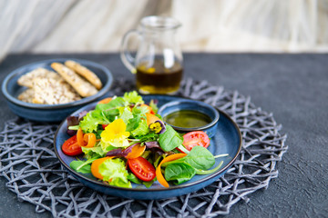 Fresh vegetable salad with tomatoes, carrot slices, greens, flowers, olive oil with herbs, balsamic cream, blue plate, dark textured table. Healthy, diet, vegan, organic food concept. Close up view.