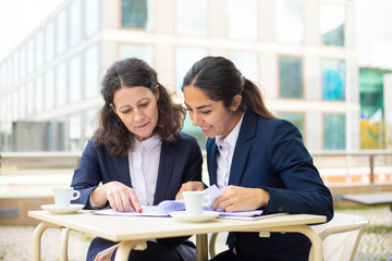 Businesswomen working with documents in outdoor cafe. Professional female colleagues in formal wear sitting at table, drinking coffee and discussing work in outdoor cafe. Coffee time concept