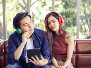  young Asian couple sitting close together on couch ,wearing headphones  listening to music from tablet.