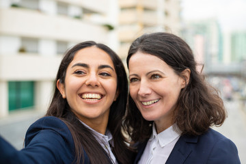 Beautiful happy businesswomen. Attractive cheerful female colleagues in formal wear talking selfie and looking aside on urban city street. Partnership concept