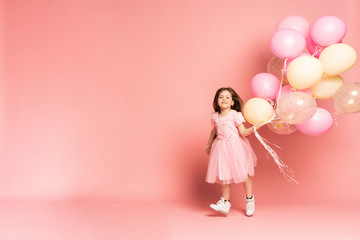 Happy celebration of birthday party with flying balloons of charming cute little girl in tulle dress smiling to camera isolated on pink background. Charming smile, expressing happiness